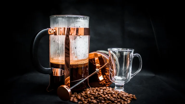 French press beside a glass cup with coffee beans scattered around it